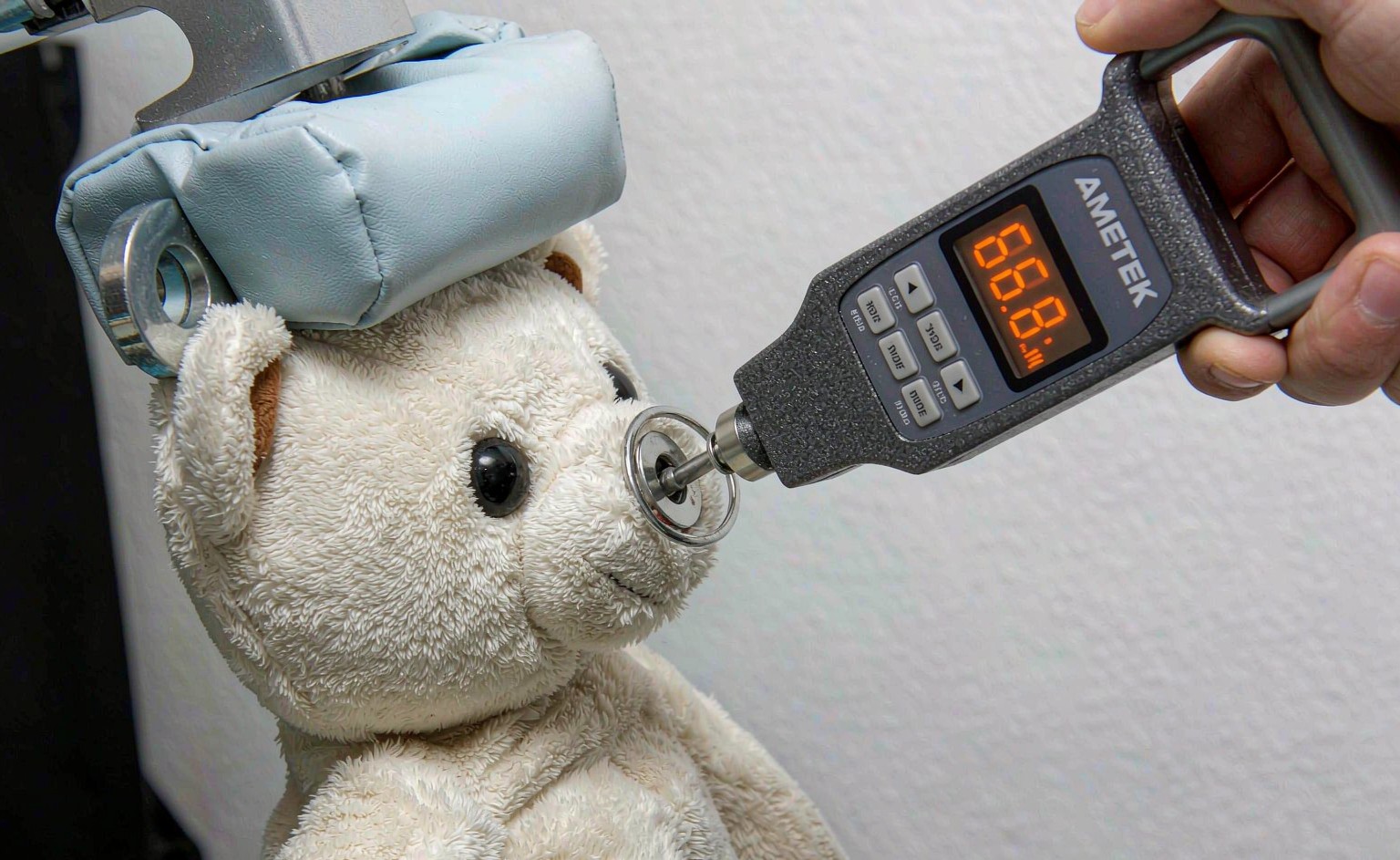 Close-up photograph of a plush toy eye being pulled with a digital force gauge during ASTM F963 tension testing, showing the 66.8N force reading on the display