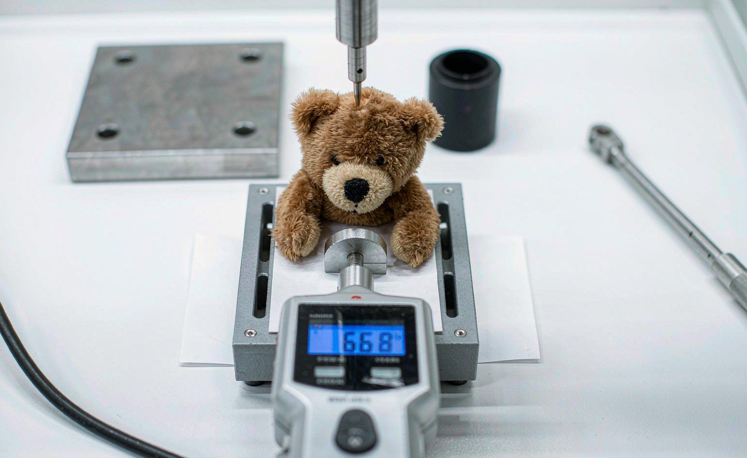 A laboratory testing technician performing ASTM F963 mechanical tests on plush toys - drop test, tension test with force gauge, and torque test setup arranged on a clean white workbench