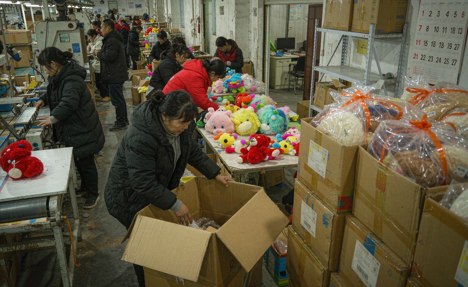 A wide documentary photograph of a busy plush toy factory floor in late January, with workers rushing to complete and pack colorful plush toy orders, finished goods in cartons stacked near the loading dock, and a calendar visible on the office wall showing the February holiday period