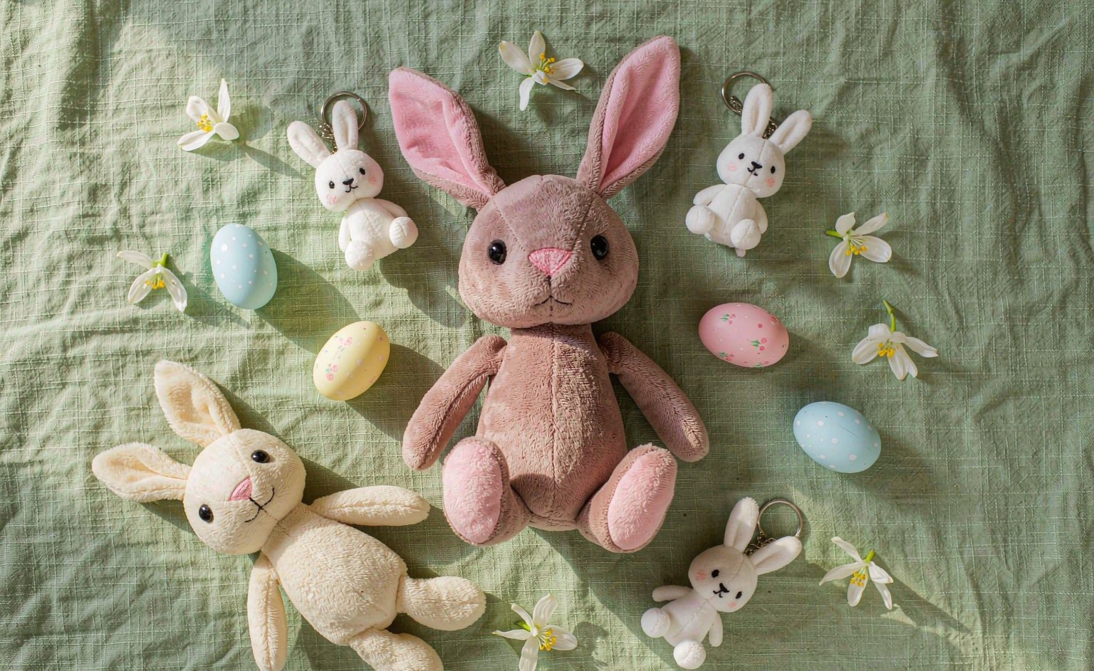 A bright spring flat-lay photograph showing an Easter 2027 plush collection: a weighted-base sitting bunny in dusty rose upright on a shelf-style surface, mini bunny keychains in pastel colors, a cream-colored evergreen bunny, and decorated Easter eggs scattered around on a soft sage-green background