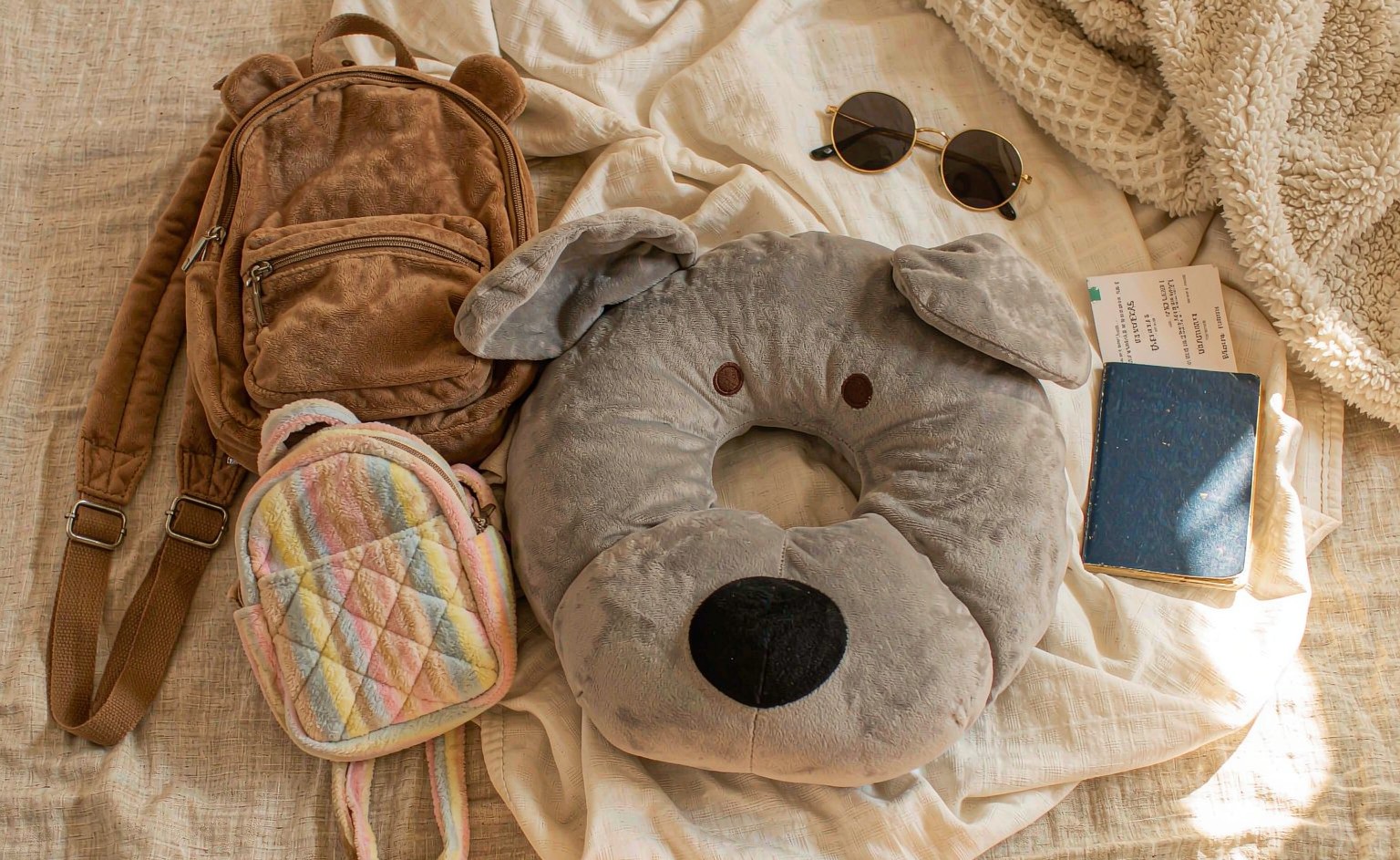A flat-lay lifestyle photograph showing functional plush products arranged on a travel-themed background: a cute animal-shaped plush travel neck pillow with memory foam core, a plush bear backpack with visible adjustable straps and zippered compartment, and a small plush crossbody bag, with a boarding pass and sunglasses nearby