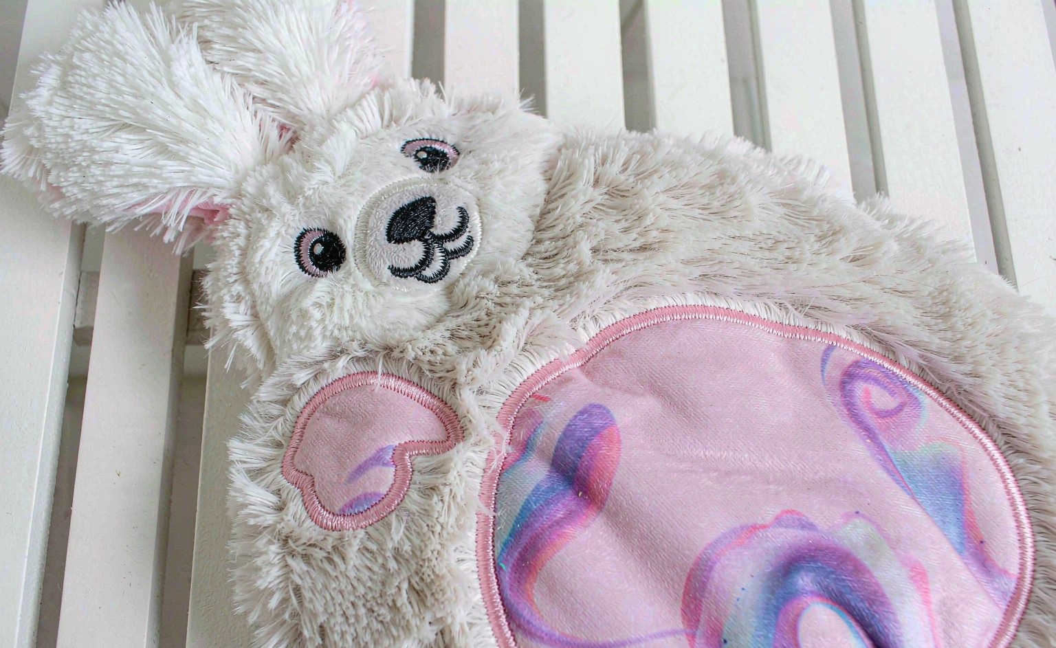 A close-up photograph of a plush toy showing hybrid decoration: crisp embroidered facial features and stitched satin-outline applique on the chest, with a colorful sublimation-printed fabric panel on its belly