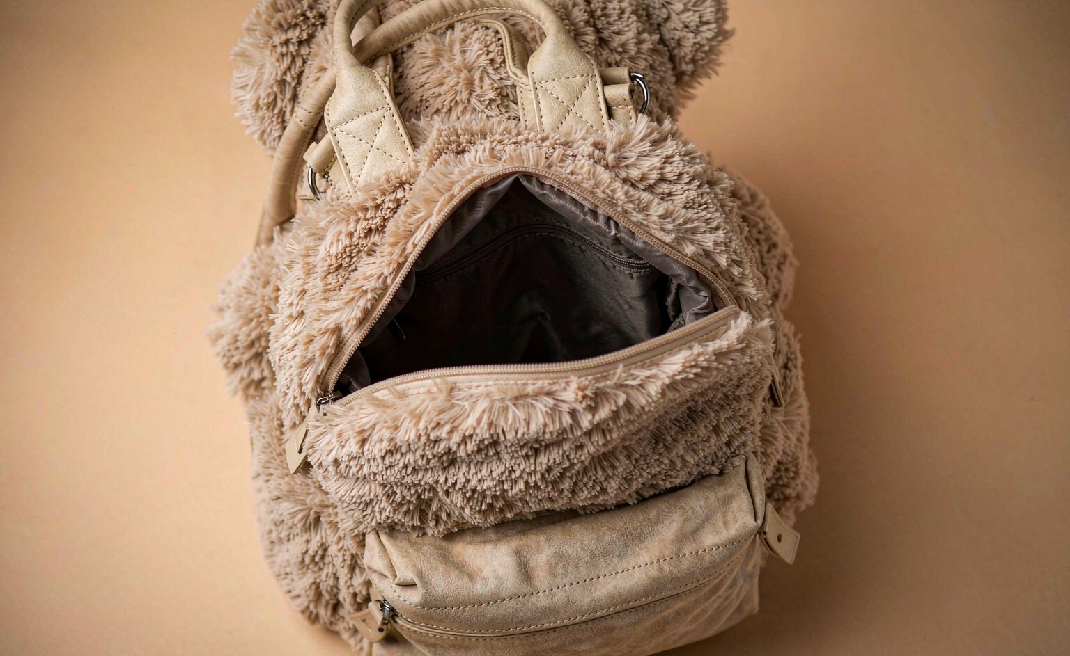 A close-up product detail photograph of a plush bear backpack showing the reinforced box-and-cross stitching on the strap anchor point, the sewn polyester lining visible through the open zippered compartment, and the structured base
