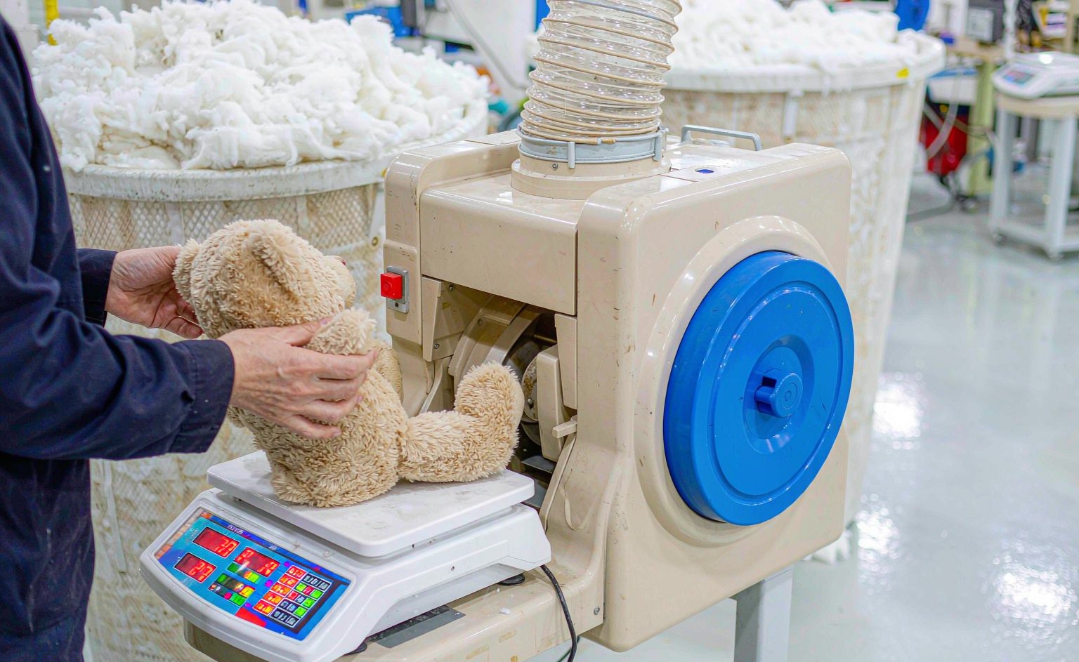 A plush factory stuffing station photograph showing a worker placing a finished plush toy on a digital weighing scale displaying its fill weight in grams, with fiber stuffing machine and bins of polyester fiberfill in the background